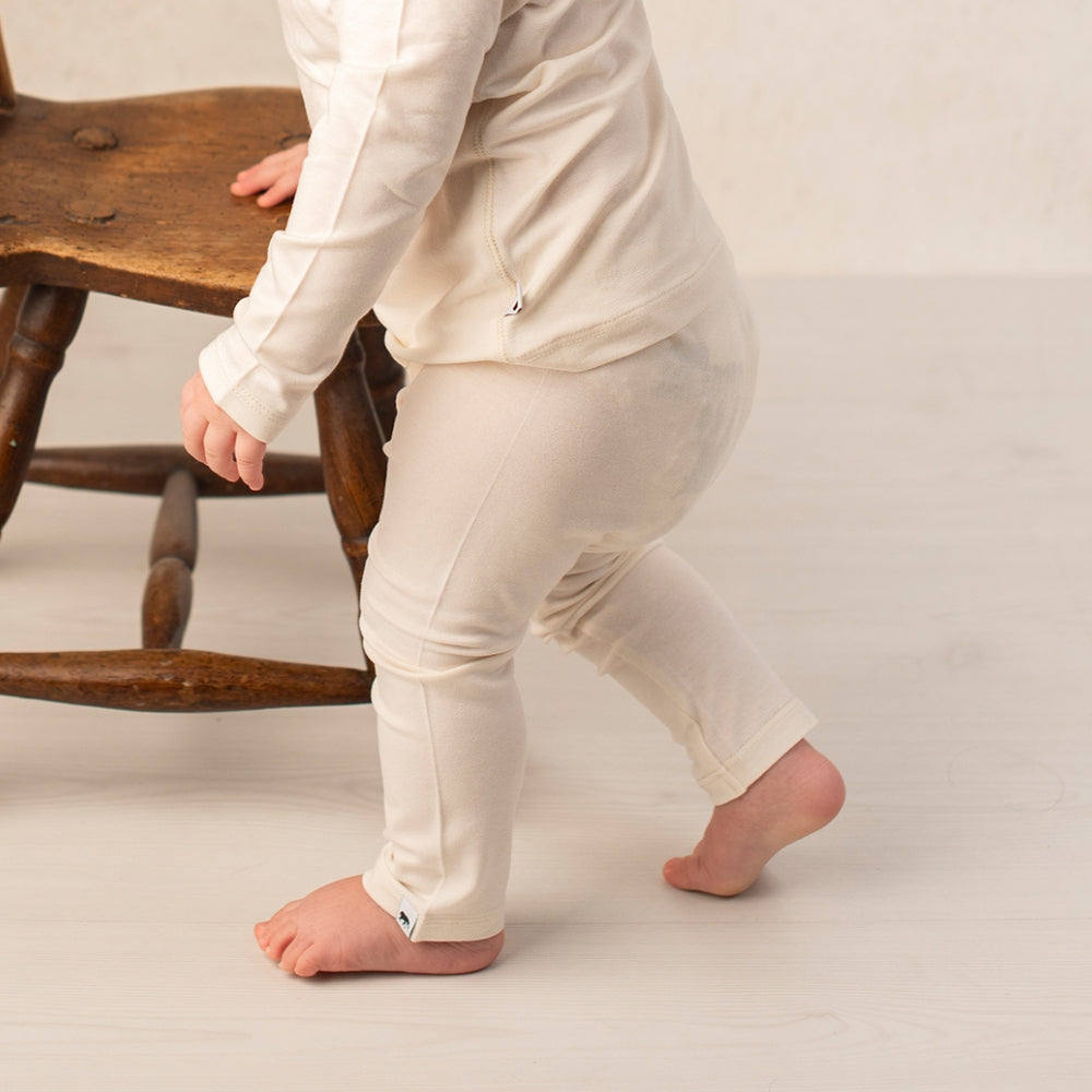 A toddler wearing a cream-colored two-piece pajama set with a long-sleeve top and matching pants, walking barefoot next to a wooden chair.