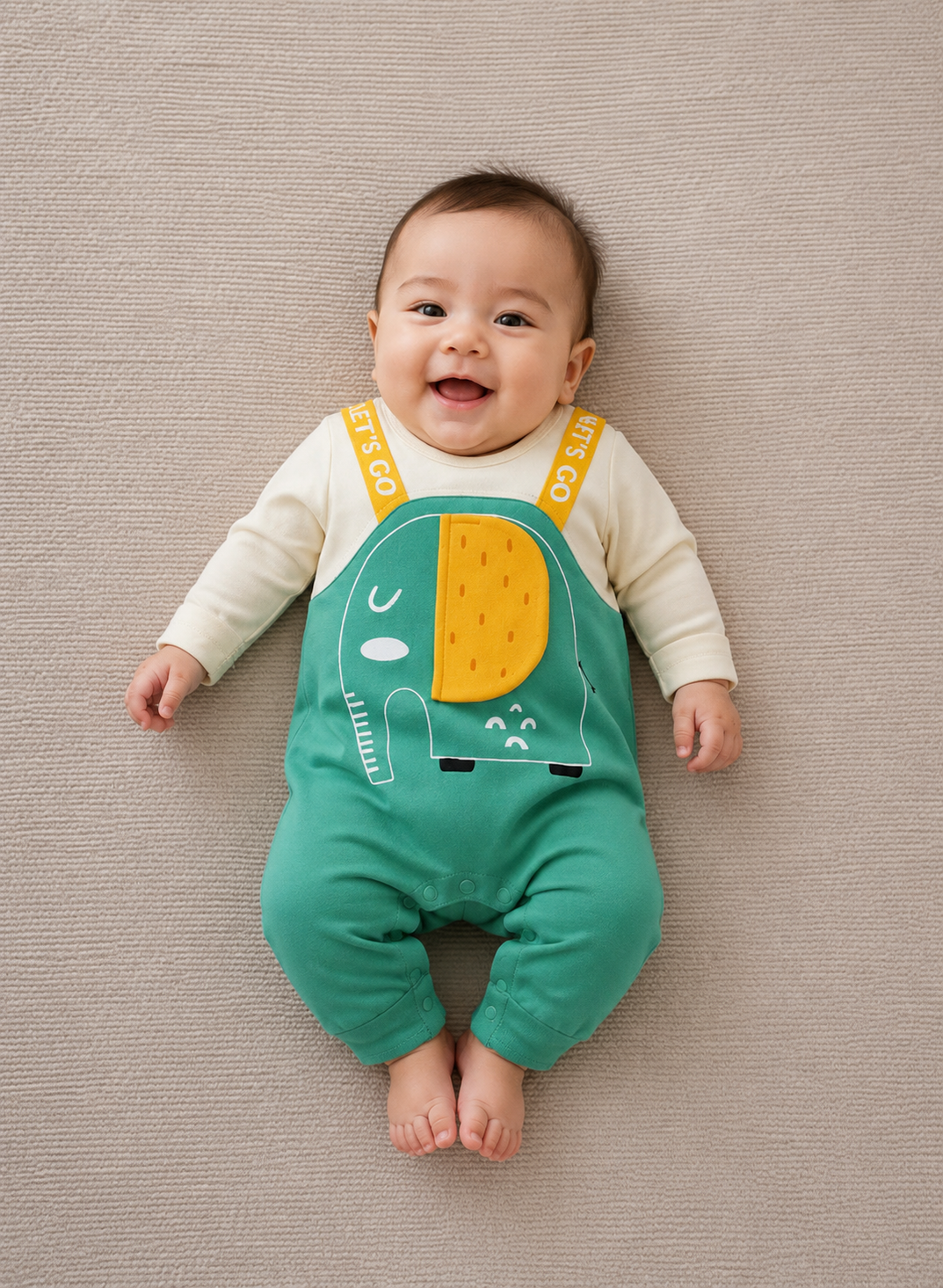 A baby lying on a textured beige blanket, wearing a cream long-sleeve top and green overall-style romper with a yellow and white elephant graphic and yellow shoulder straps printed with 'LETS GO’